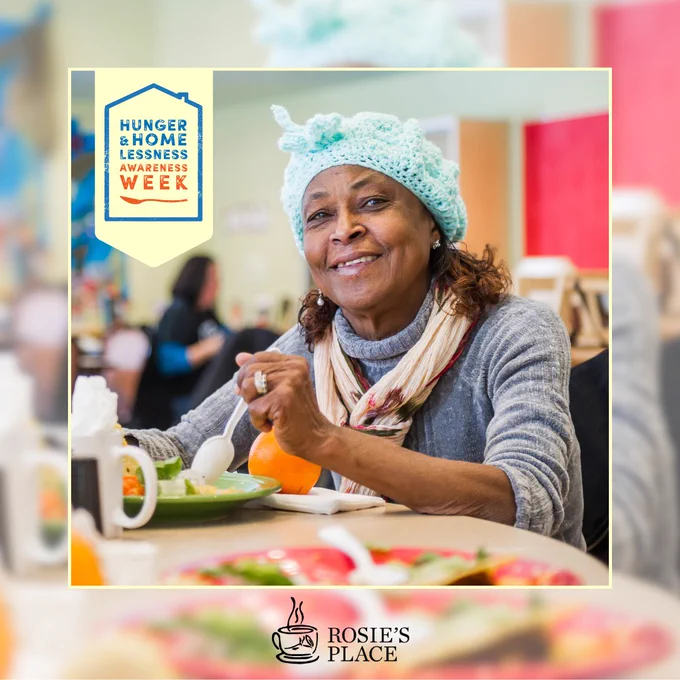 Elderly woman wearing a blue knit hat and scarf eating a meal at a community dining table with logo for Hunger & Homelessness Awareness Week.