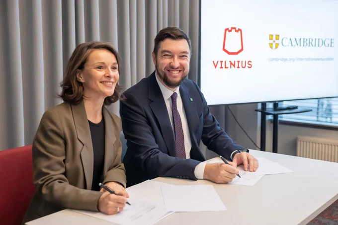 Two individuals, one is the Deputy Mayor of Vilnius City (Vytautas Mitalas), signing a Memorandum of Understanding (MoU) at a table with logos of Cambridge and Vilnius City displayed in the background. Both individuals are smiling at the camera.