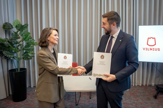Two individuals, one is the Deputy Mayor of Vilnius City, shaking hands over a table with documents, in front of a screen displaying the Vilnius city logo.