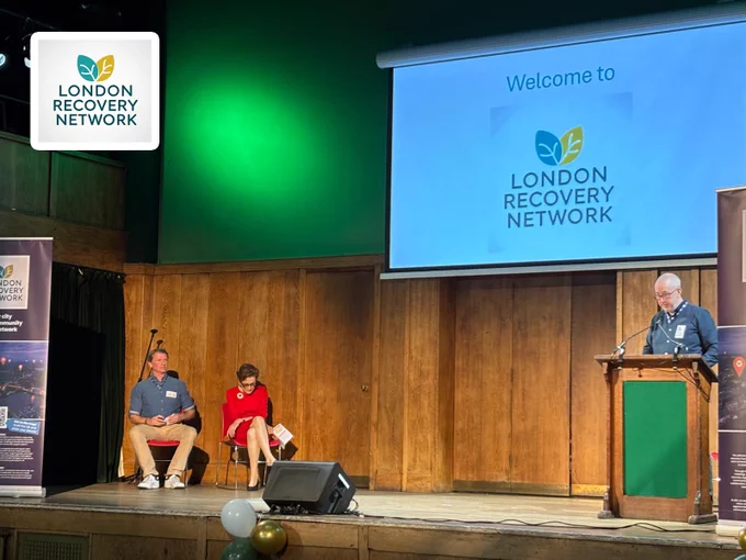 Panel discussion at London Recovery Network event with two seated participants and one speaker at podium on stage.