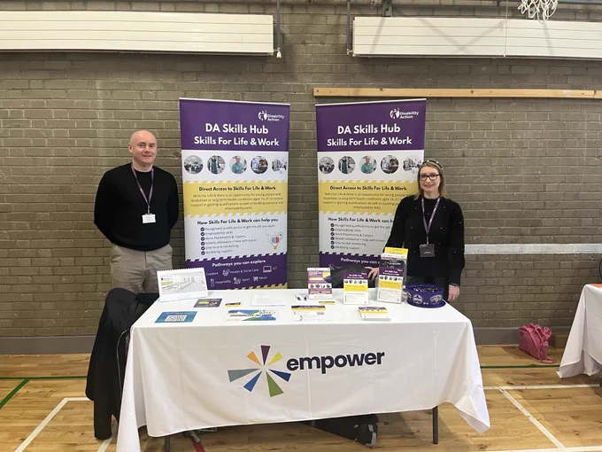 Two staff members from Disability Action stand behind a display table promoting the DA Skills Hub – Skills for Life & Work programme. The table is covered with information leaflets, flyers and purple-branded materials, with an Empower tablecloth at the front. Behind them are two large purple pop-up banners with details about the programme. They are set up in a school sports hall with brick walls and a basketball hoop visible in the background.