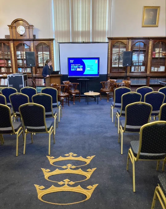 A person stands beside a screen displaying "Staff Briefing May 2023" in a traditional library hall with rows of empty blue chairs, wooden bookshelves, and a large crest on the carpet.