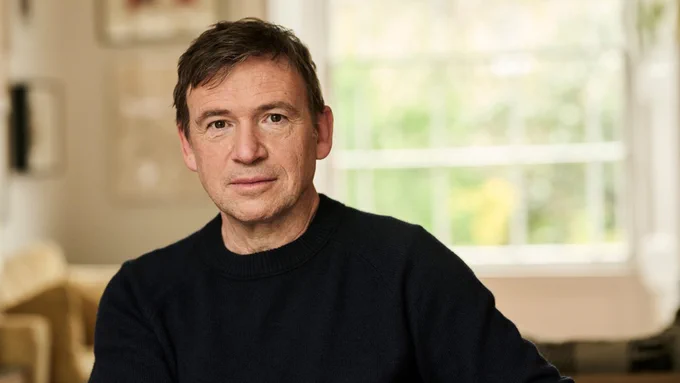 A man in a dark jumper smiles to camera, sitting in a brightly lit room.