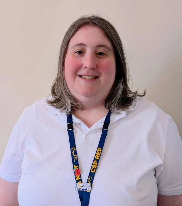 Photo of Christina smiling at the camera. Christina is wearing a white shirt and a lanyard, and is stood in front of a beige wall.