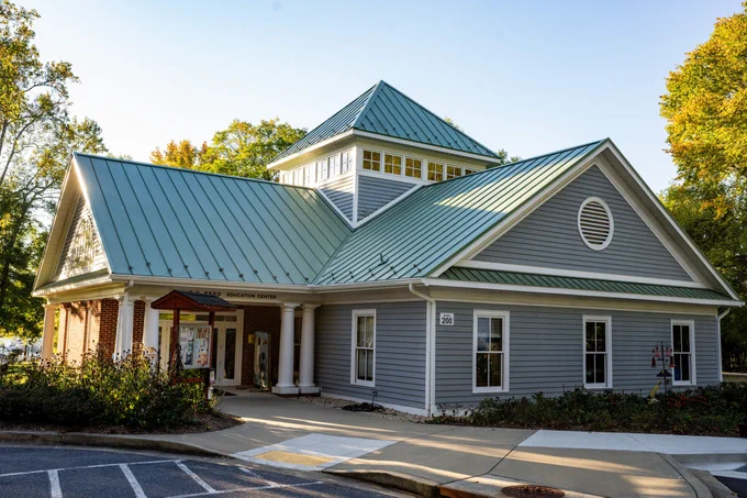 An L-shaped building with gray siding and a light green roof sits beneath a blue sky, surrounded by trees. The words "Reed Education Center" are above the doorway.