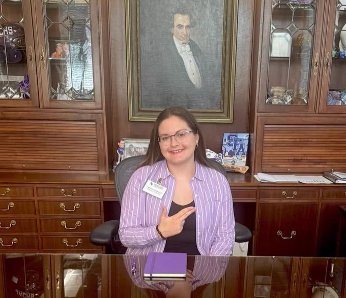 Person smiling, sitting at an office desk with a book, in front of a portrait of Stephen F Austin
