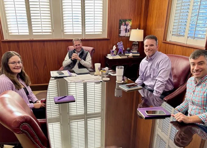 Four people sitting around a conference table, smiling and looking at the camera, in a wood-paneled room with diplomas on the wall. 