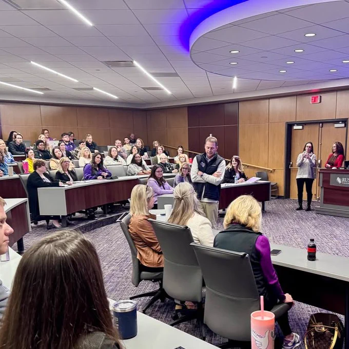 A conference room filled with attendees listening to a speaker. The room features modern decor and purple accent lighting.