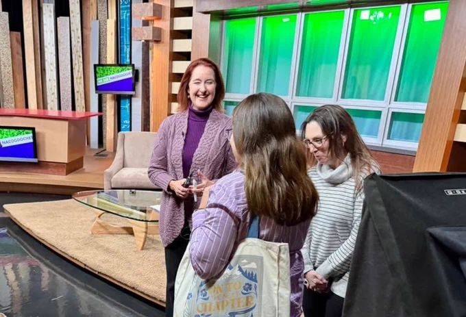 Four individuals on a television studio set, three engaged in conversation with smiles, and one looking towards the camera equipment. The setting includes modern decor and a professional broadcasting atmosphere.