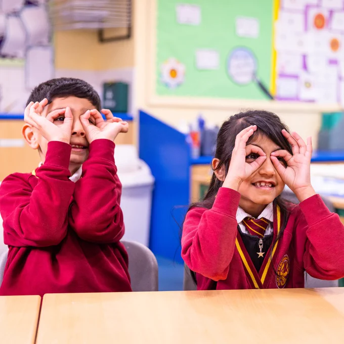A girl and a boy, about 9-years-old, wear red school uniforms as they sit in a classroom. They use their fingers to make the shape of glasses over their eyes, as if to see all the better.