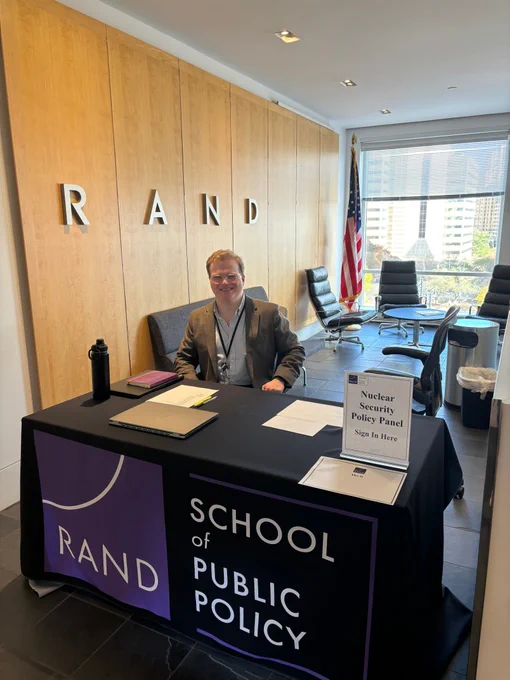A man is smiling behind a RAND School of Public Policy table, upon which is a sign that reads "Nuclear Security Policy Panel"