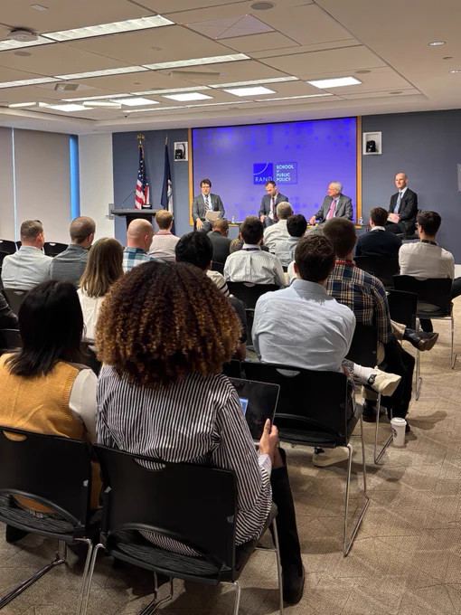 At the front of a conference room, four men in suits are seated in front of a RAND School of Public Policy screen and an audience of people. 