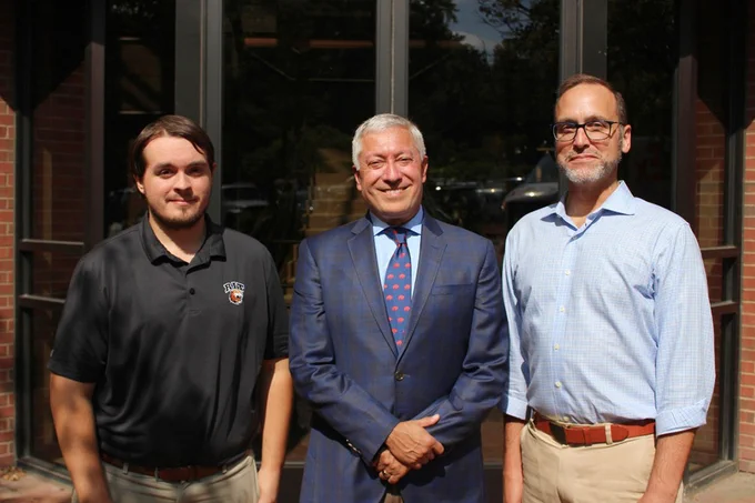 Three men pose for a picture. On the left is a student with short brown hair wearing a black polo shirt. Center is an older man with white hair wearing a blue suit and tie. On the right is a middle-aged man with short brown hair and a short grey beard wearing glasses and a button-down shirt.