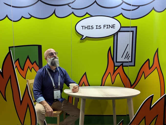 Bearded guy sitting in the "this is fine" set patterned after the meme, complete with flames all around. He's staring off into space, holding a cup of coffee. Things are definitely not fine. 