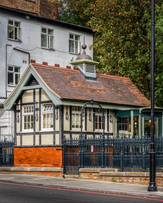 A photograph of a building with Tudor-style architecture, featuring exposed timber framing, white walls, and a shingled roof. Surrounded by ornate black iron fencing.