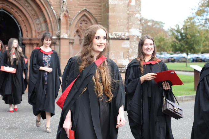 Graduates in black robes and red stoles smiling as they walk away from a historic university building.