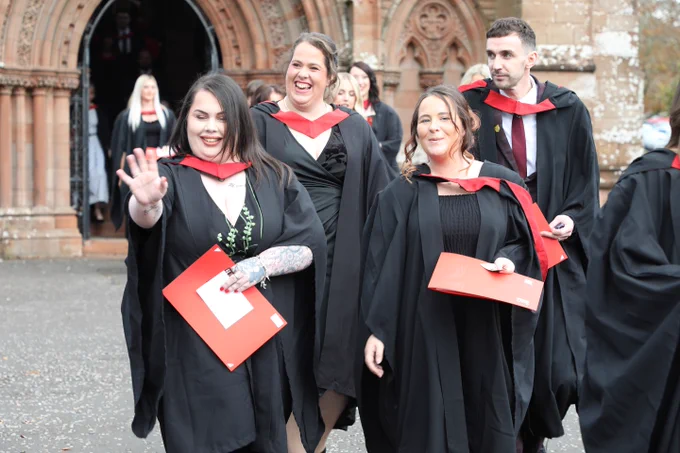 Group of graduates in caps and gowns, smiling and walking away from a historic building, holding their diploma folders.