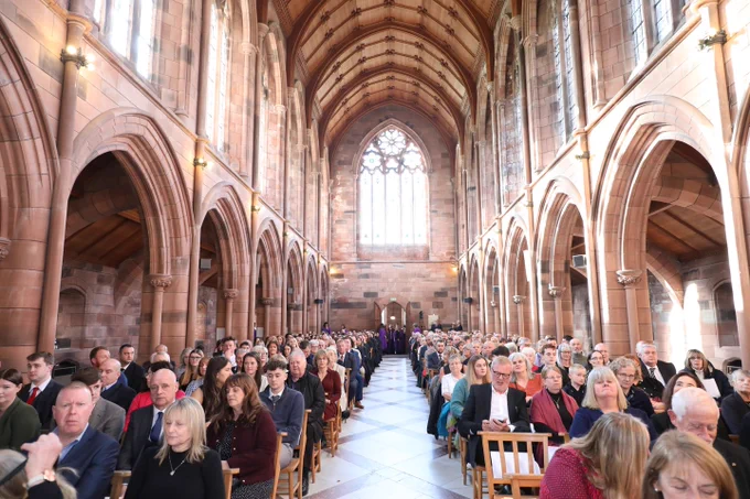 Interior of a cathedral with vaulted ceilings and stained glass, filled with attendees seated in rows during an event.