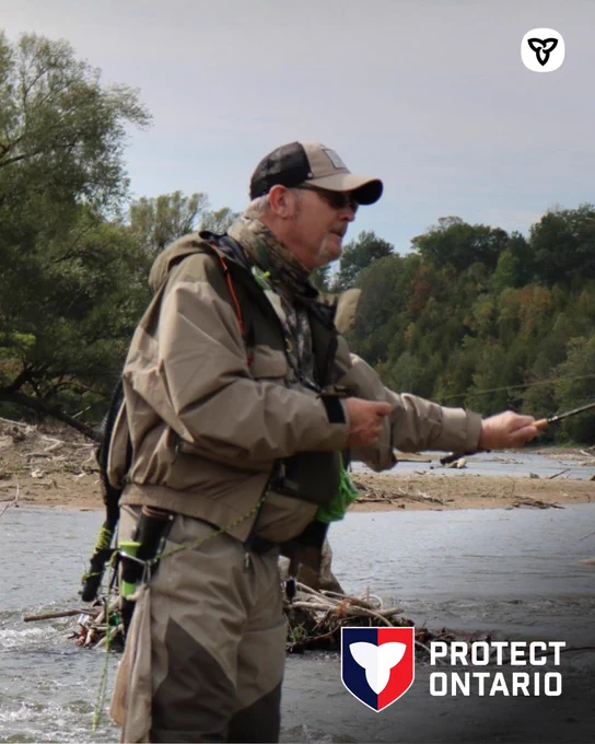 Man in tan jacket, pants and hat fishing in a river. 
