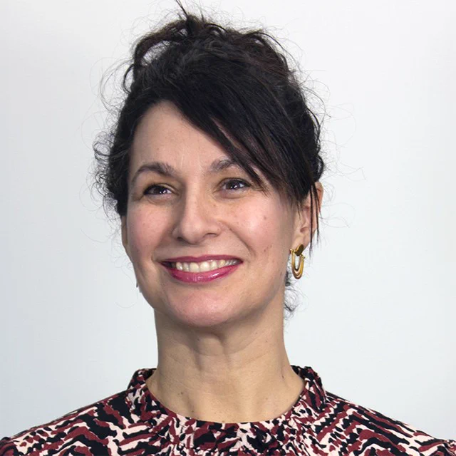 A woman with dark curly hair tied up wearing gold hoop earrings and a patterned red, black, and white top against a plain light background.