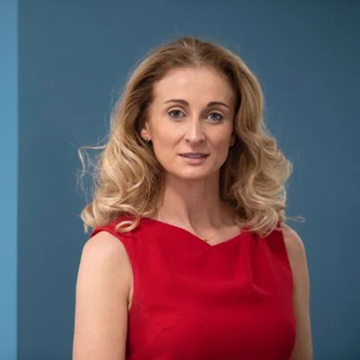 Woman with shoulder-length blonde curly hair wearing a sleeveless red dress against a blue background.
