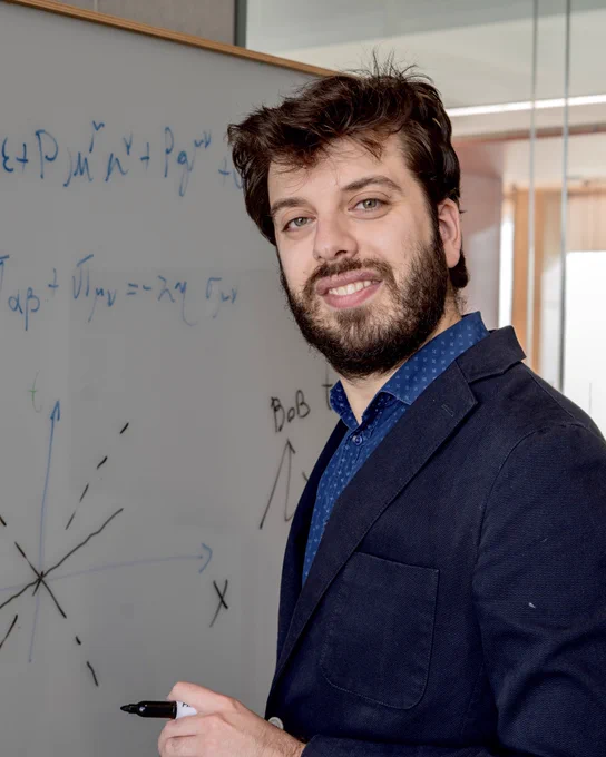 Physicist Lorenzo Gavassino, recipient of the 2026 APS Valley Prize, stands beside a whiteboard filled with equations and diagrams, holding a marker and smiling toward the camera.