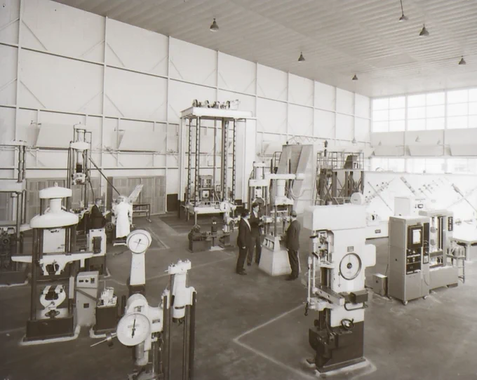 Spacious industrial workshop with vintage machinery and two workers inspecting equipment under high ceilings and large windows.