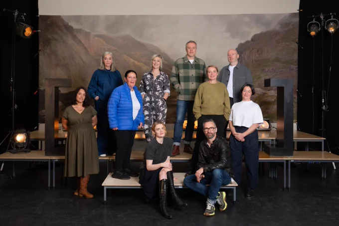 A group of ten people smiling together on a stage in front of a painted backdrop of mountains. Some are standing on platforms, while two are seated in front. 