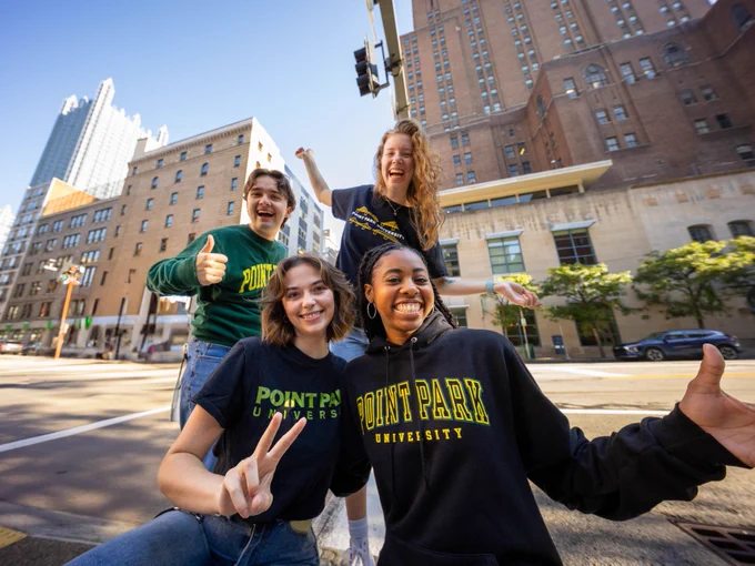 Four students wearing Point Park University sweatshirts smiling and posing outdoors on a sunny city street, making cheerful gestures.
