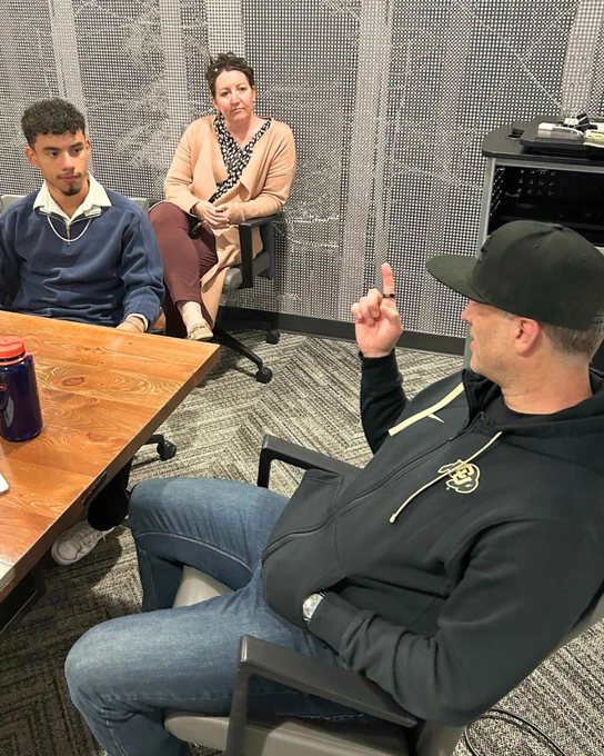 Three individuals sitting around a table in a meeting room, one person is speaking and gesturing with their hand.
