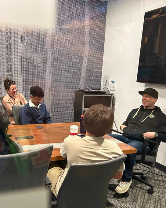 Group of people in a casual meeting with an adult in a black ballcap seated around a table in a room with a glass wall.