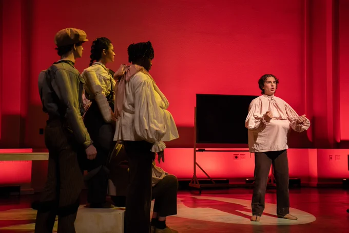 Performers on stage in a theatrical production with a red backdrop. One individual stands in the foreground speaking, while three others listen, dressed in vintage costumes.