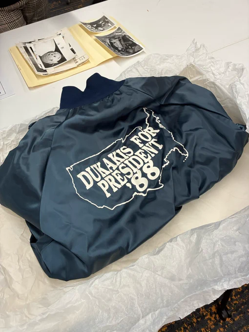 A dark blue jacket sits folded on a table in white tissue paper. The jacket has an outline of the continental United States with "DUKAKIS FOR PRESIDENT '88" inside