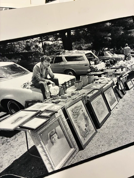 Black and white image of a man sitting on the hood of an old car. In front of him are a line of tables covered with framed artwork. More artwork leans against the front of the tables