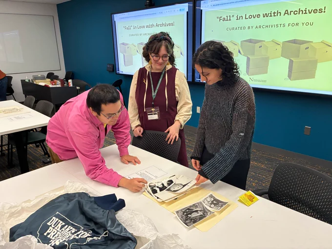 Three people look at archival materials on a table. One is pointing to an image while the other two look on
