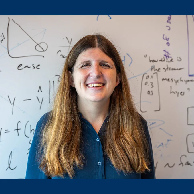 Emma Pierson stands and smiles in front of a whiteboard with mathematical equations.