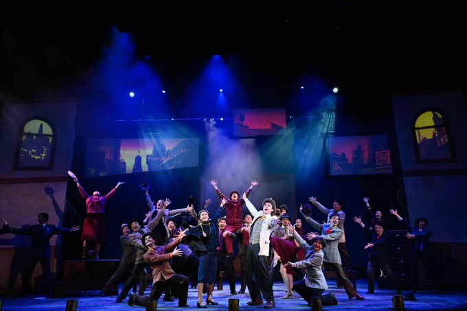 A vibrant stage scene from the musical "Guys and Dolls" with performers energetically singing and gesturing under atmospheric blue stage lighting.