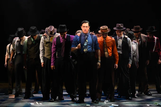 A person stands prominently in the center on stage surrounded by a group wearing hats, lit by dramatic stage lighting, during a scene from a theatrical production.