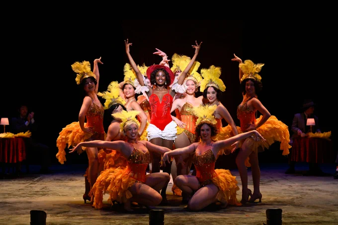 Performers in vibrant cabaret costumes with feathers on stage, centered around a lead singer in a red and gold outfit, during a lively show.
