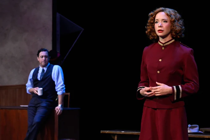 Two actors performing on stage in a theater setting, with one standing in the background holding a cup and another in the foreground dressed in a red outfit.