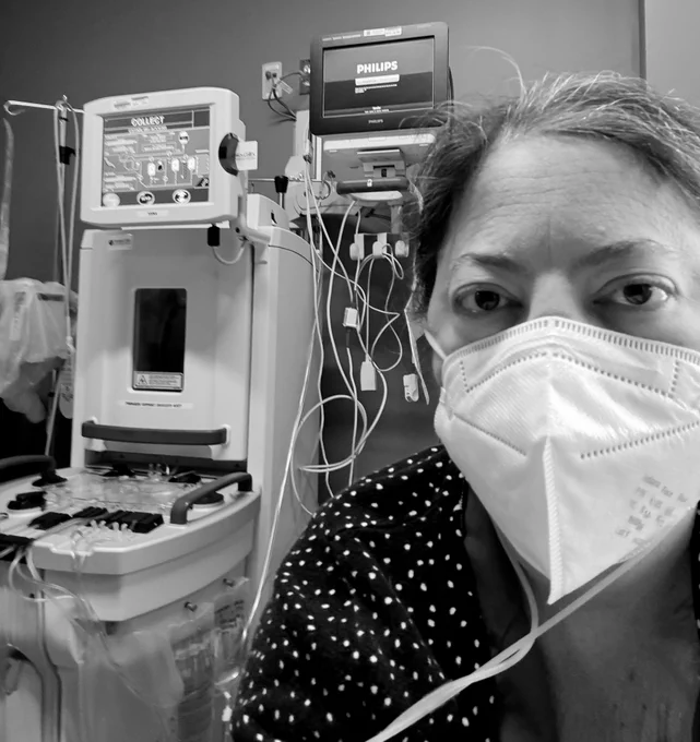 Black and white photo in medical clinic of middle aged white woman with dark hair pulled back looking straight ahead wearing an oxygen cannula under N95 mask and a white polka dot sweater.  To the left in the background is the pheresis machine.
