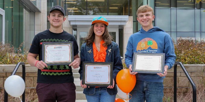 Three students holding achievement certificates stand in front of a university building, with two wearing university-branded apparel.