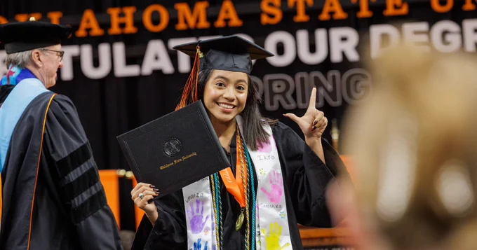 A joyful graduate at Oklahoma State University holds a diploma and gestures a peace sign, wearing a cap and gown adorned with colorful stoles.