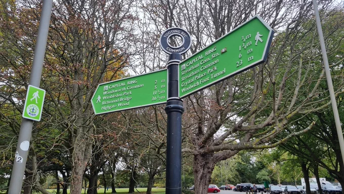 An iconic lime green London Capital Ring fingerpost just outside Tooting Bec Lido.  The backdrop to the post is autumnal trees and parkland, a distant green field and carpark.