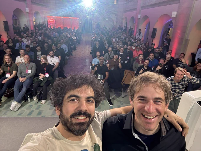 Two speakers smile for a selfie on stage in front of a large, cheerful audience seated in rows inside a bright hall with arched walls and pink lighting. The crowd, many wearing conference badges, waves and makes peace signs toward the camera. A strong light beam shines from the back of the room, creating a radiant atmosphere.