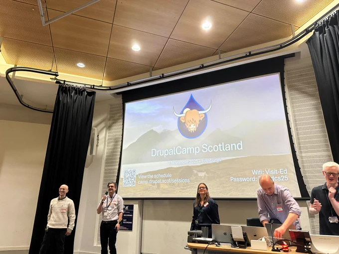 Photo of DrupalCamp Scotland stage, showing multiple presenters along with the DrupalCamp Scotland logo, a stylised highland cow superimposed on the Drupal drop logo.