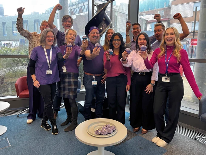 A group of researchers and clinical trialists in a research building, wearing purple, holding purple cupcakes and cheering.
