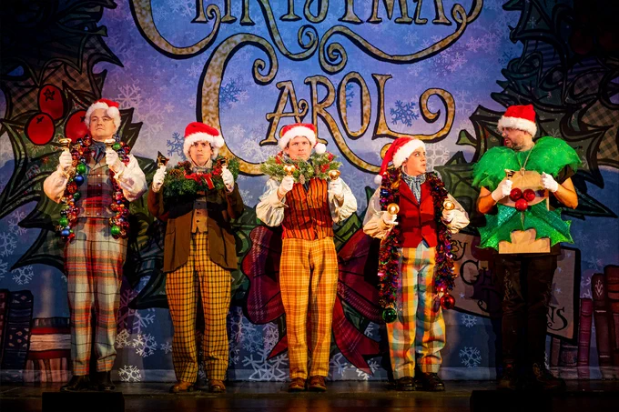 A scene from Christmas Carol Goes Wrong featuring five actors on stage in festive, Dickensian costumes