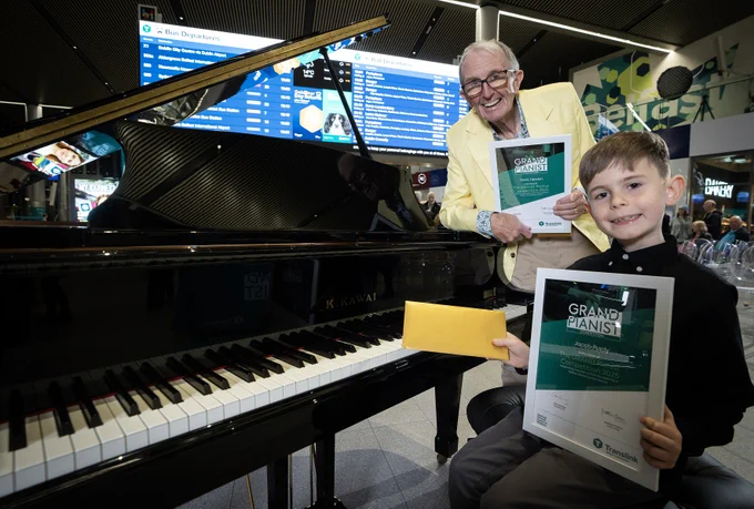 A black grand piano is positioned in an indoor public space with a large digital departures board visible in the background. Two people are standing and sitting near the piano, each holding certificates titled “Grand Pianist” and a yellow envelope. The setting appears to be an event or award presentation at a transport hub, with modern architecture and signage visible behind them.