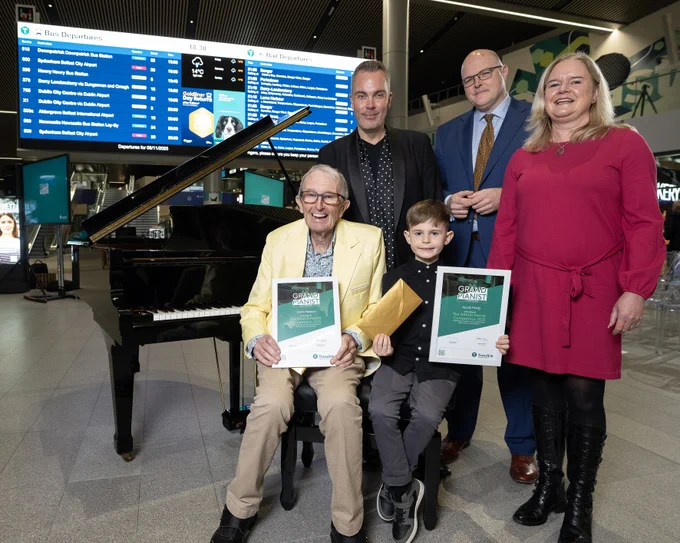 “Group photo taken in a modern transport hub with a grand piano in the foreground. Two seated individuals hold certificates, while three others stand behind them. A large digital departures board and promotional screens are visible in the background.
